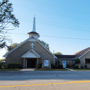 Multistory tan brick church building with white steeple with cross on top and parking lot