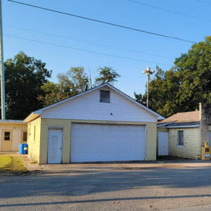 one yellow concrete block building with wide white garage door and small blond brick building with legs of water tower behind