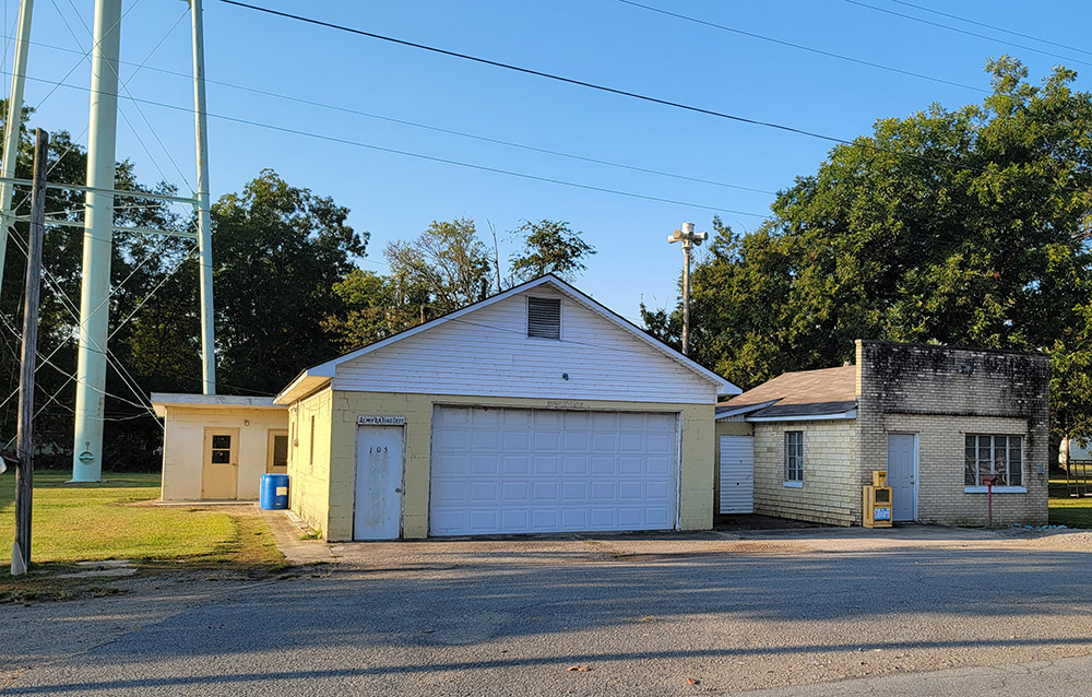 one yellow concrete block building with wide white garage door and small blond brick building with legs of water tower behind