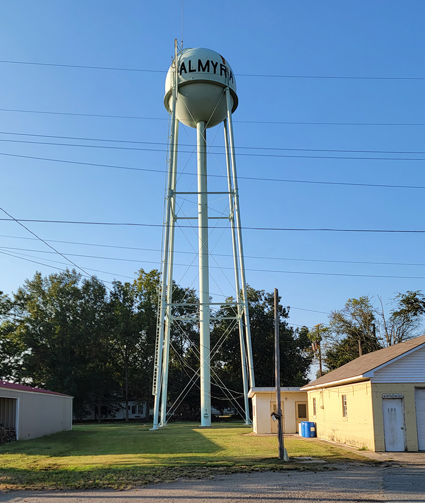Metal cylindrical water tank on tall legs with "Almyra" in large black letters