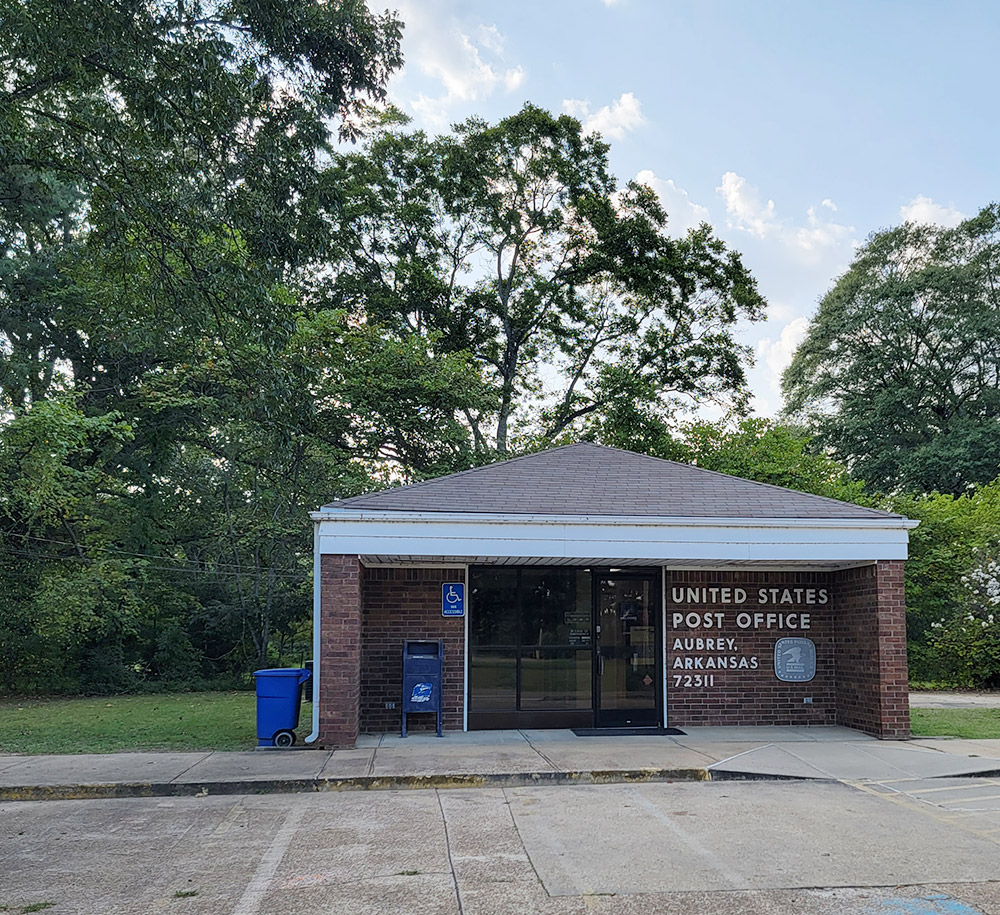 Single story red brick post office building with parking lot and trees in background