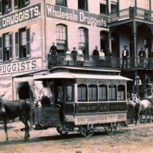 Horse drawn rail cars on city street with an audience standing on balcony of multistory buildings