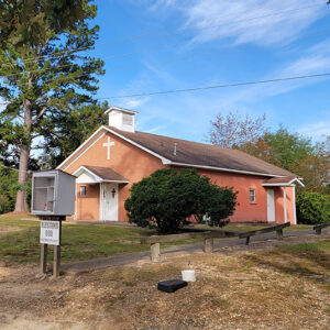 Single story red brick church building with trees and a "blessing box" pantry in front