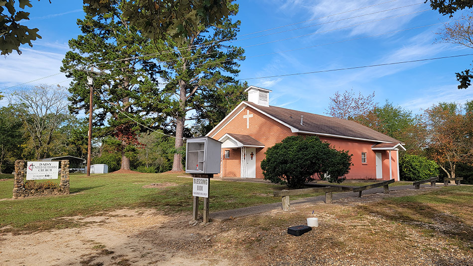 Single story red brick church building with trees and a "blessing box" pantry in front