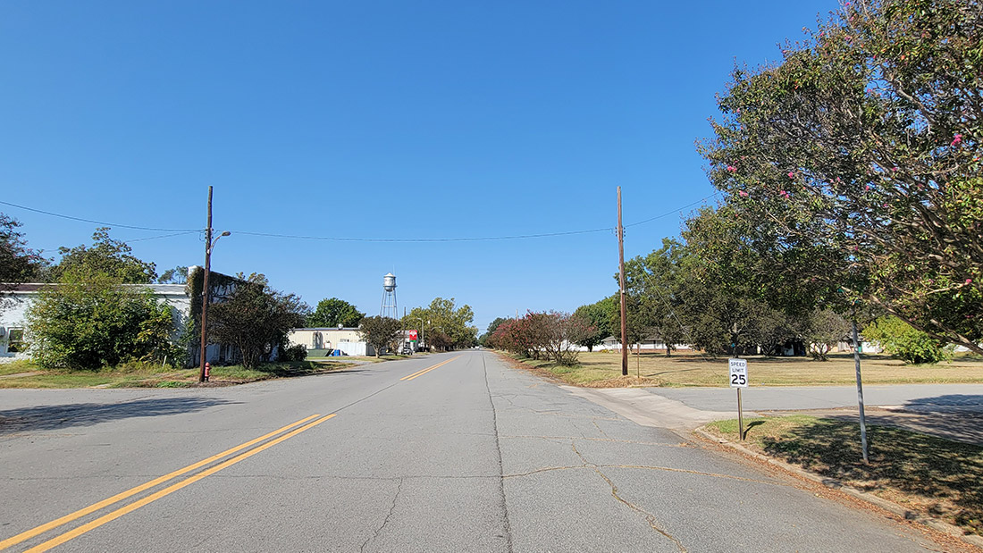 Small town street scene with low buildings obscured by trees and a water tower in the distance