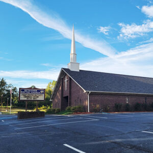 Single story red brick church building with steeple and parking lot