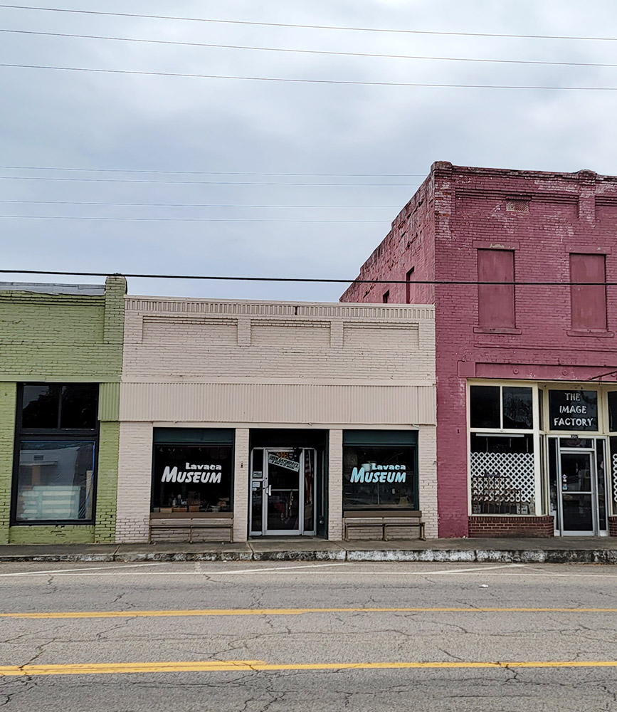Single story beige brick building in row of storefronts with "Lavaca Museum" written on both front windows