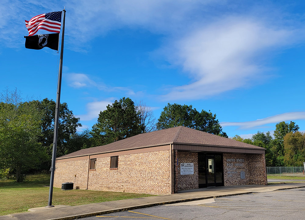 Single story blond brick post office building with flagpole flying American and P.O.W. flags