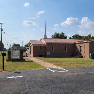 Single story red brick church building with red roof and white steeple and parking lot
