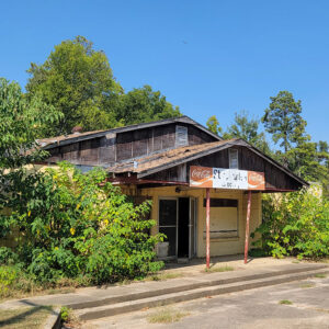 dilapidated single story wooden grocery store building with Coke signs and partly covered by vegetation