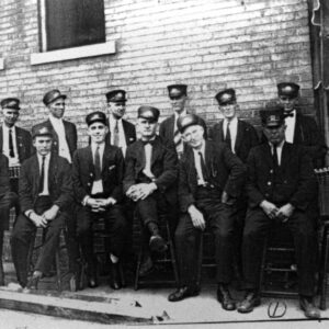 Seated group of white men in streetcar conductor uniforms and hats next to building wall