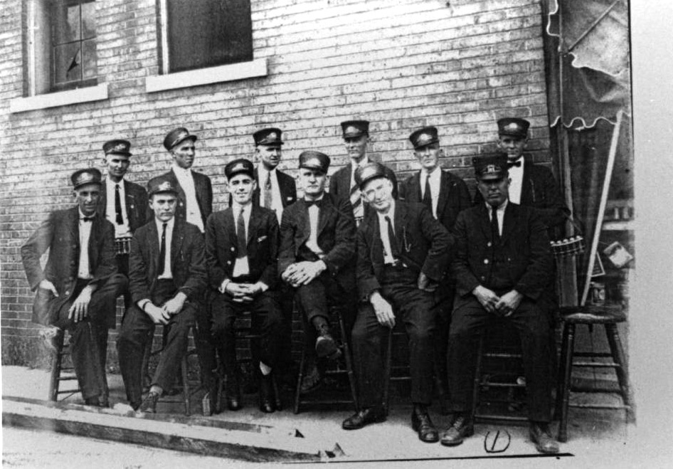 Seated group of white men in streetcar conductor uniforms and hats next to building wall