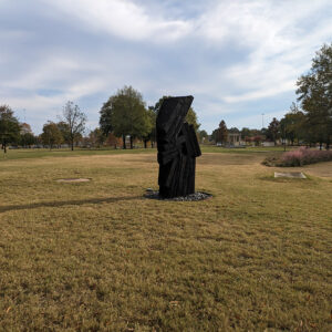An abstract sculpture sitting in a large grassy place with trees and a gazebo in the distance