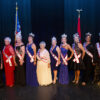 Eight white and two black older women dressed in evening wear and crowns and sashes with state and American flags in background