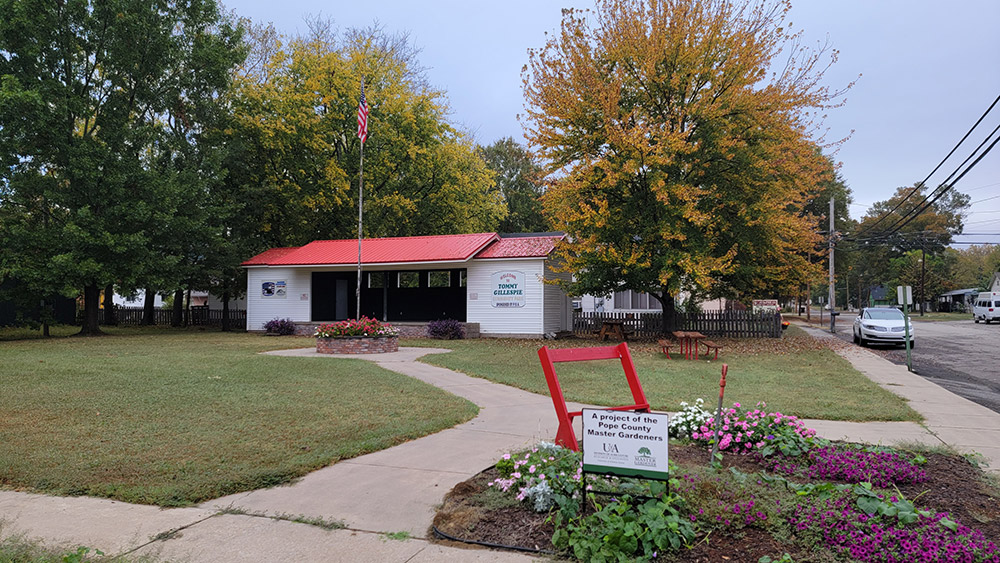 Entrance to a city park with small white building with red roof with trees around and a garden display in front