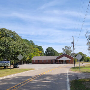 Single story orange brick church building with parking lot and trees on curving highway