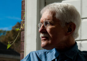 side view of a white man with white hair and glasses staring into the distance