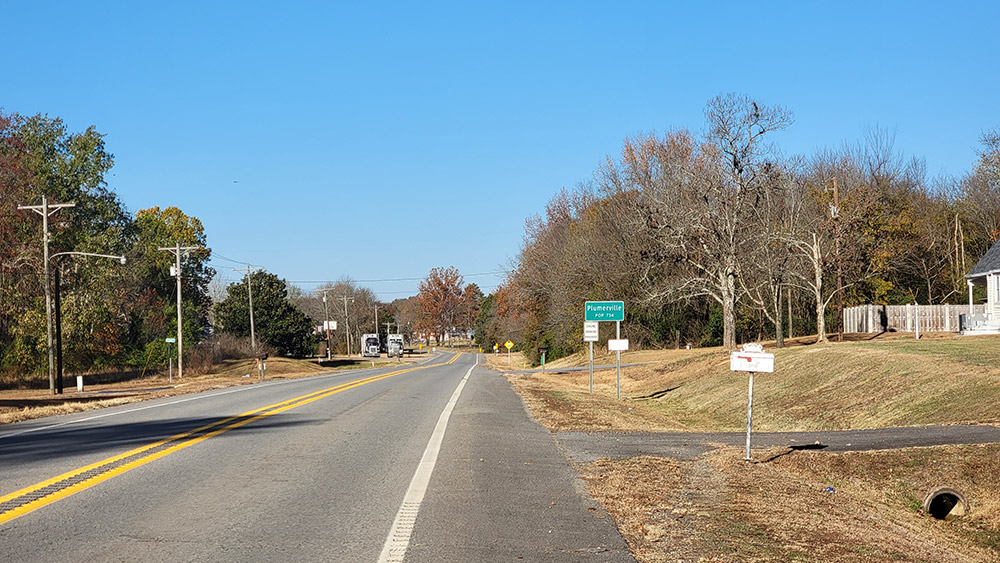 Highway with two trucks and trees on both sides and sign saying "Plumerville"