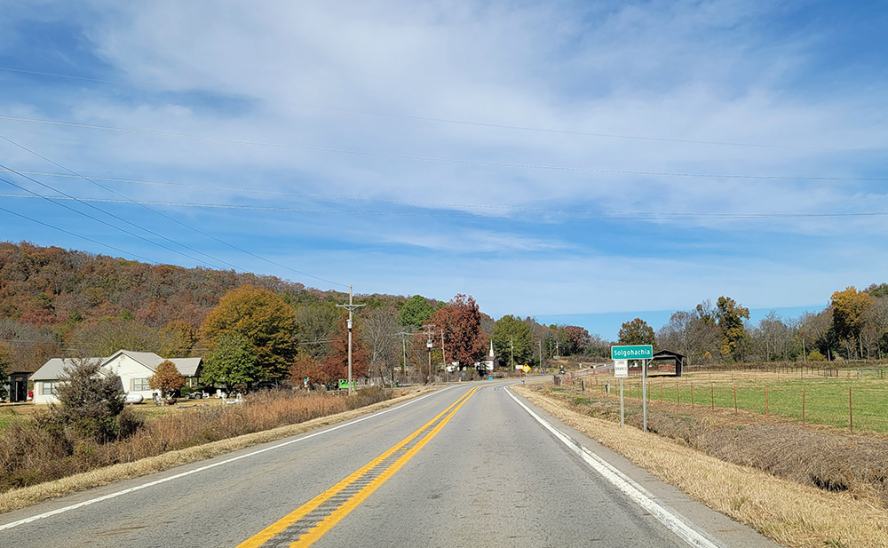 Country road entering a small town with tree-lined hill in distance on left
