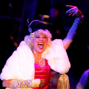 Female actor in fancy dress and hat on stage with her hand up and her mouth open