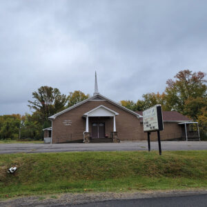 Single story brown brick church building with white steeple and parking lot