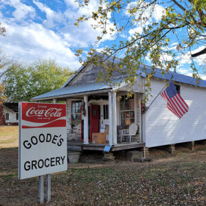 Single story white wooden grocery store building with covered entrance and sign "Goode's Grocery"