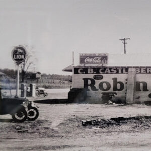Single story white wooden building with old cars and gas pumps and "C. B. Castleberry Grocery" and "Robin Hood" in big letters on the side