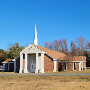 Single story orange brick church building with white columns and a steeple with a gazebo beside