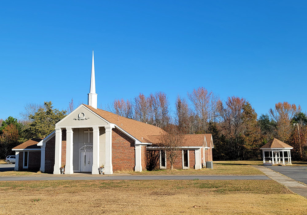 Single story orange brick church building with white columns and a steeple with a gazebo beside