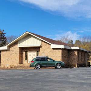 Single story tan brick church building with parking lot and green car parked in front
