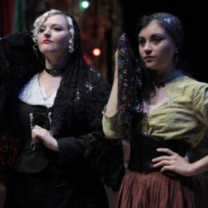 Two white women on stage holding ornate scarves up to their faces