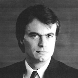 White man with brown hair wearing suit and tie and small button