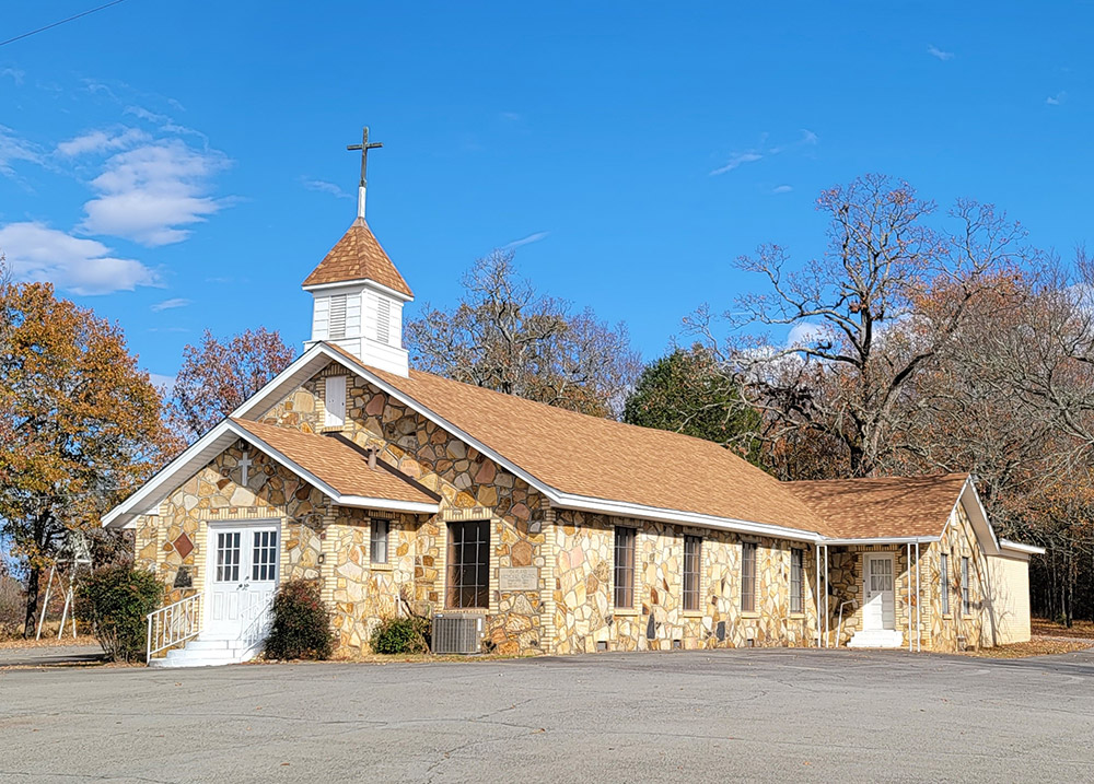Single story rock church building with small steeple with cross on top and parking lot