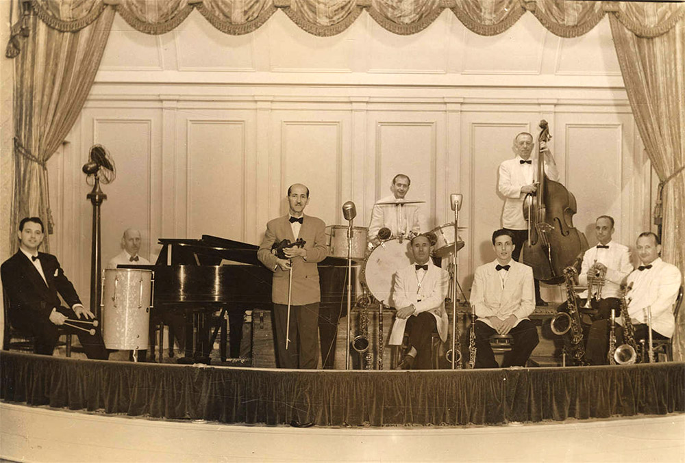 Nine white musicians in white jackets and black bow ties holding various musical instruments