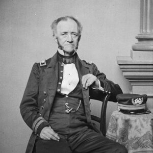 White man in naval military garb sitting on chair with cap on nearby table