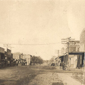 Old photo of street scene in small town dirt road lined with storefronts and horse-drawn carriages