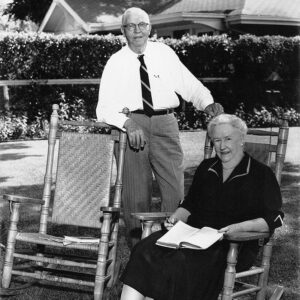 White man in a tie standing and white woman in a dress seated in rocking chair