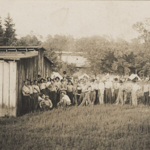 a couple dozen white men standing outside a wooden building with trees in background