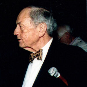 side view of a white man in a suit and bow tie holding a cane under his arm