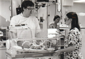 White man and white woman examining baby in incubator