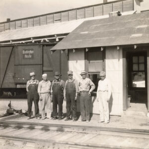 Six men standing outside at a rail yard. Behind them are two buildings and a train car with tracks in the foreground